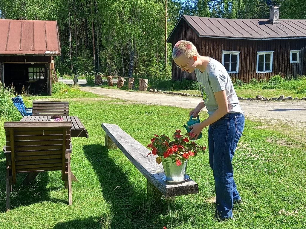 Nuori nainen kastelee kukkia maalaistalon pihapiirissä.