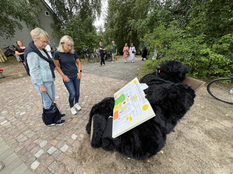 Kaksi naista katselee täytettyä karhua vanhan pihapiirin edessä.