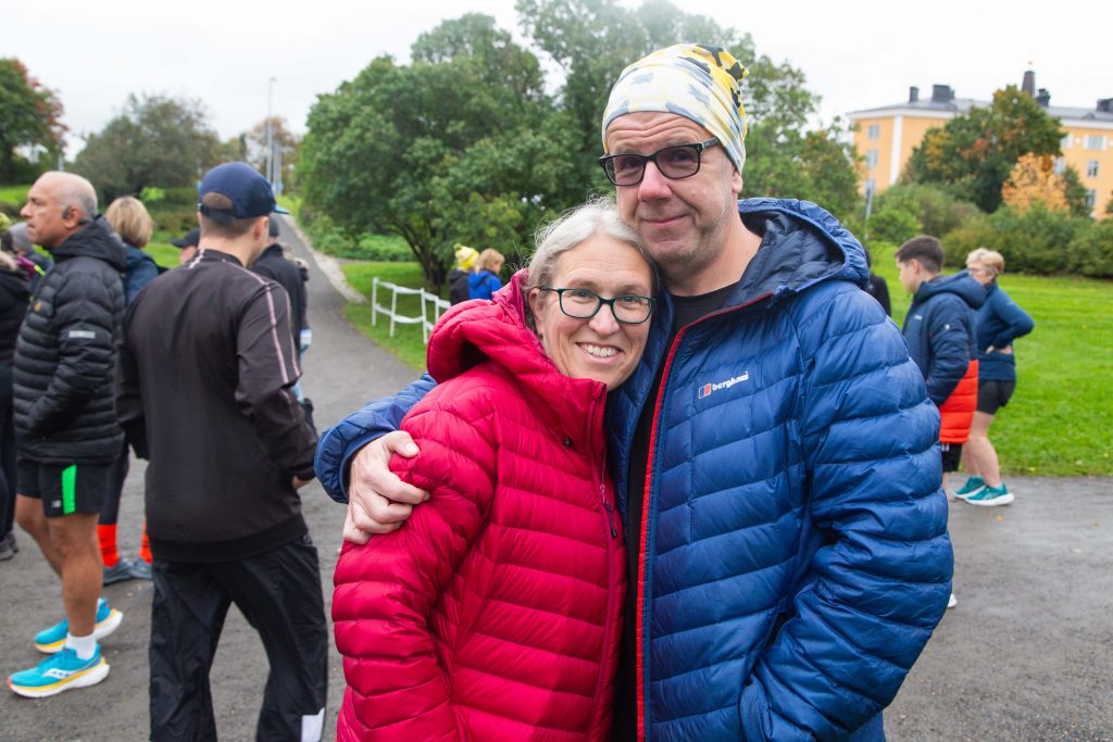 Pariskunta poseeraa puistossa, taustalla muita kuntoilijoita.