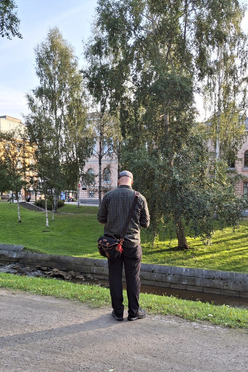 MIes seisoo puistokäytävällä sekin kameraan. Hänen takanaan näkyy nurmikkoa ja puita sekä vanha kivitalo.