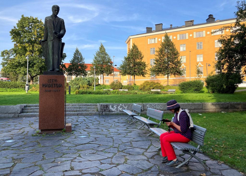 Nainen istuu kuvan oikeassa laidassa puistonpenkillä ja tutkii matkapuhelintaan. Kuvan vasemmassa laidassa olevan patsaan hahmo kuin katsoo naista jalustaltaan.