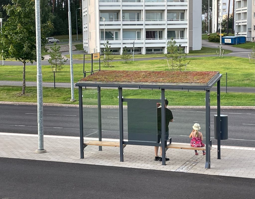 Ihmisiä bussipysäkillä, jossa viherkatos.