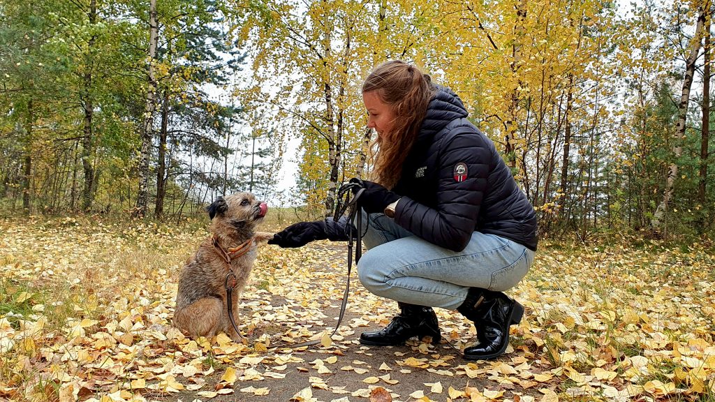 Nainen kyykyssä koiran kanssa syksyisessä maisemassa.