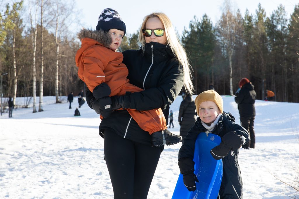 Äiti ja kaksi lasta pulkkamäen edustalla, toinen lapsi sylissä, toinen pitelee pulkkaa.